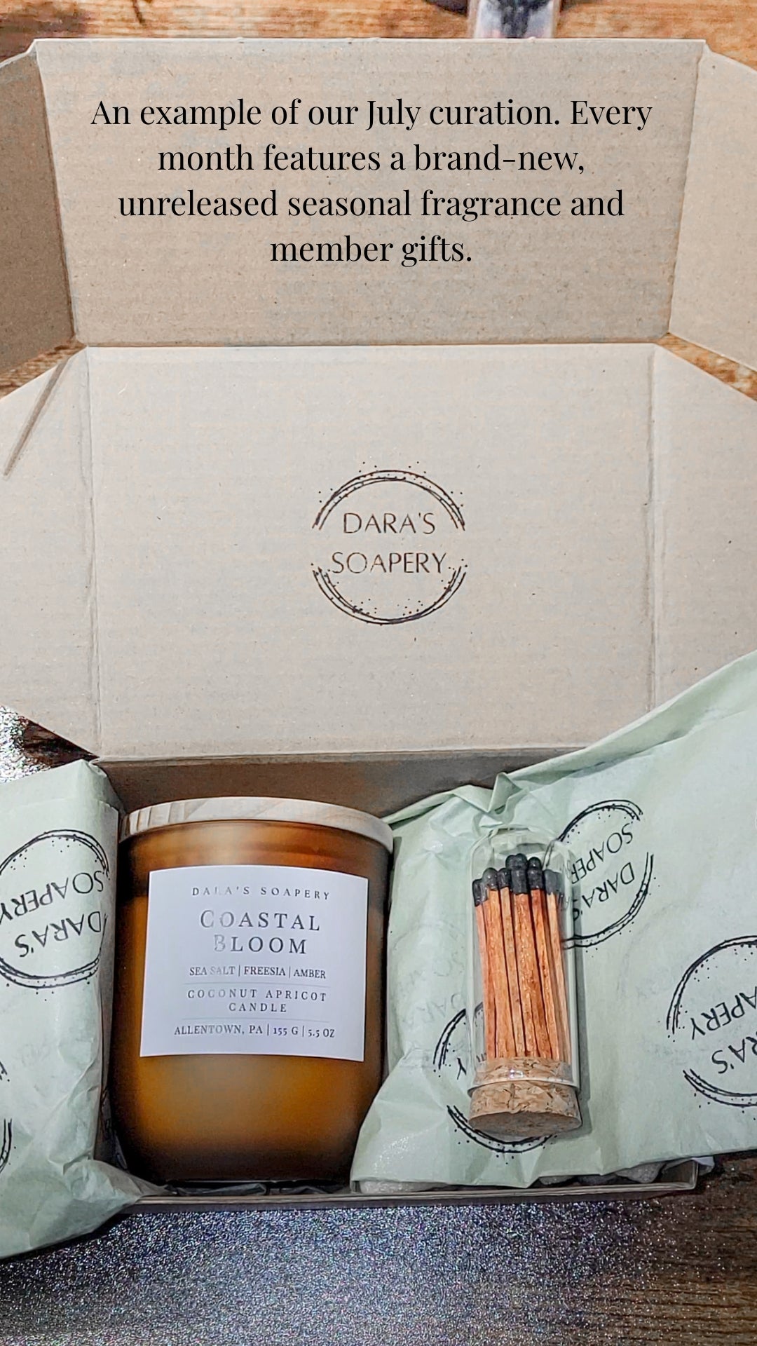 An open cardboard gift box from Dara's Soapery containing a jar candle labeled Coastal Bloom, a wrapped soap or gift item, and a small matchstick container, with printed text at the top describing monthly fragrance and member gifts.