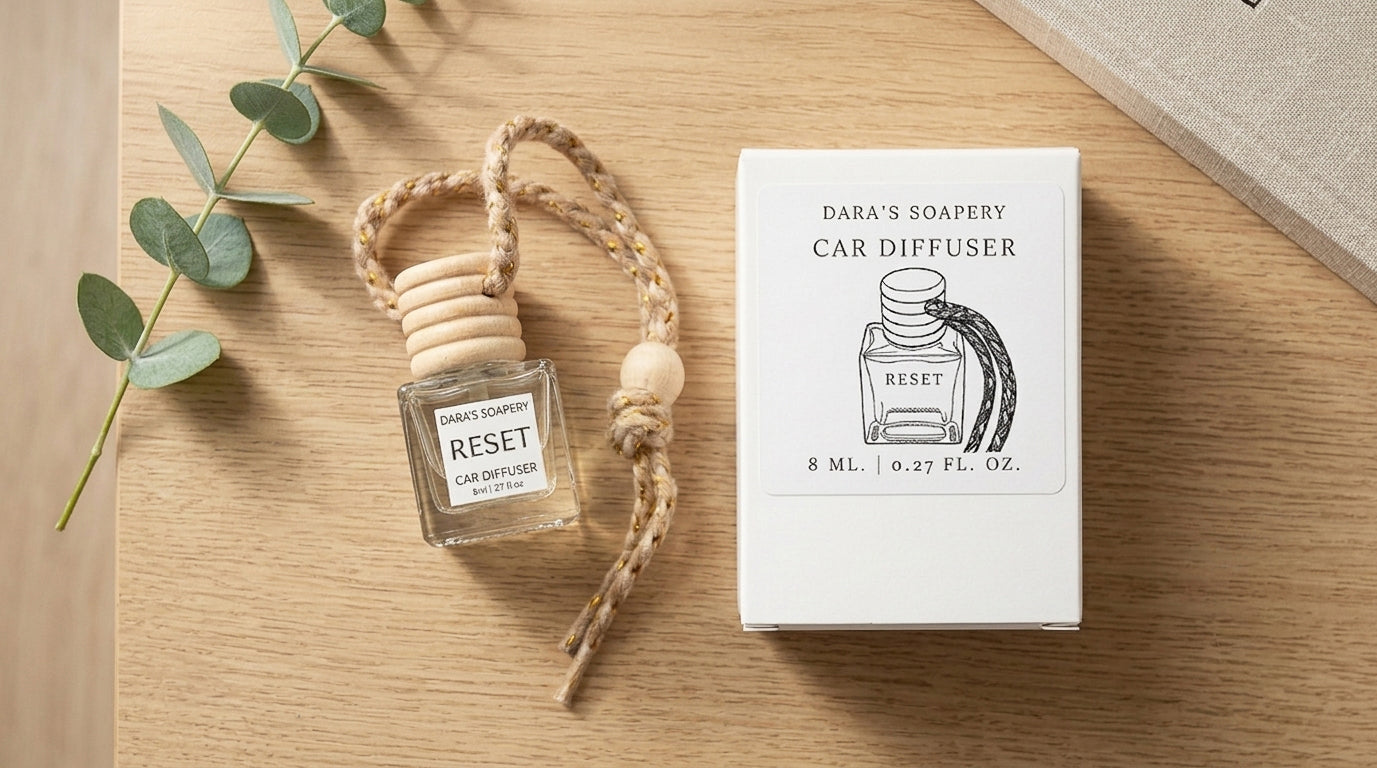 A Dara's Soapery car diffuser bottle with a wooden top and rope loop, shown next to its white product box on a light wood surface with eucalyptus leaves and a book edge as props.