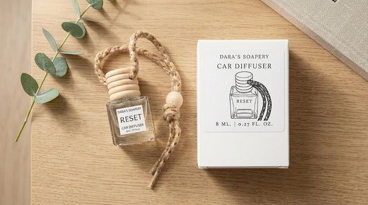 A Dara's Soapery car diffuser bottle with a wooden top and rope loop, shown next to its white product box on a light wood surface with eucalyptus leaves and a book edge as props.