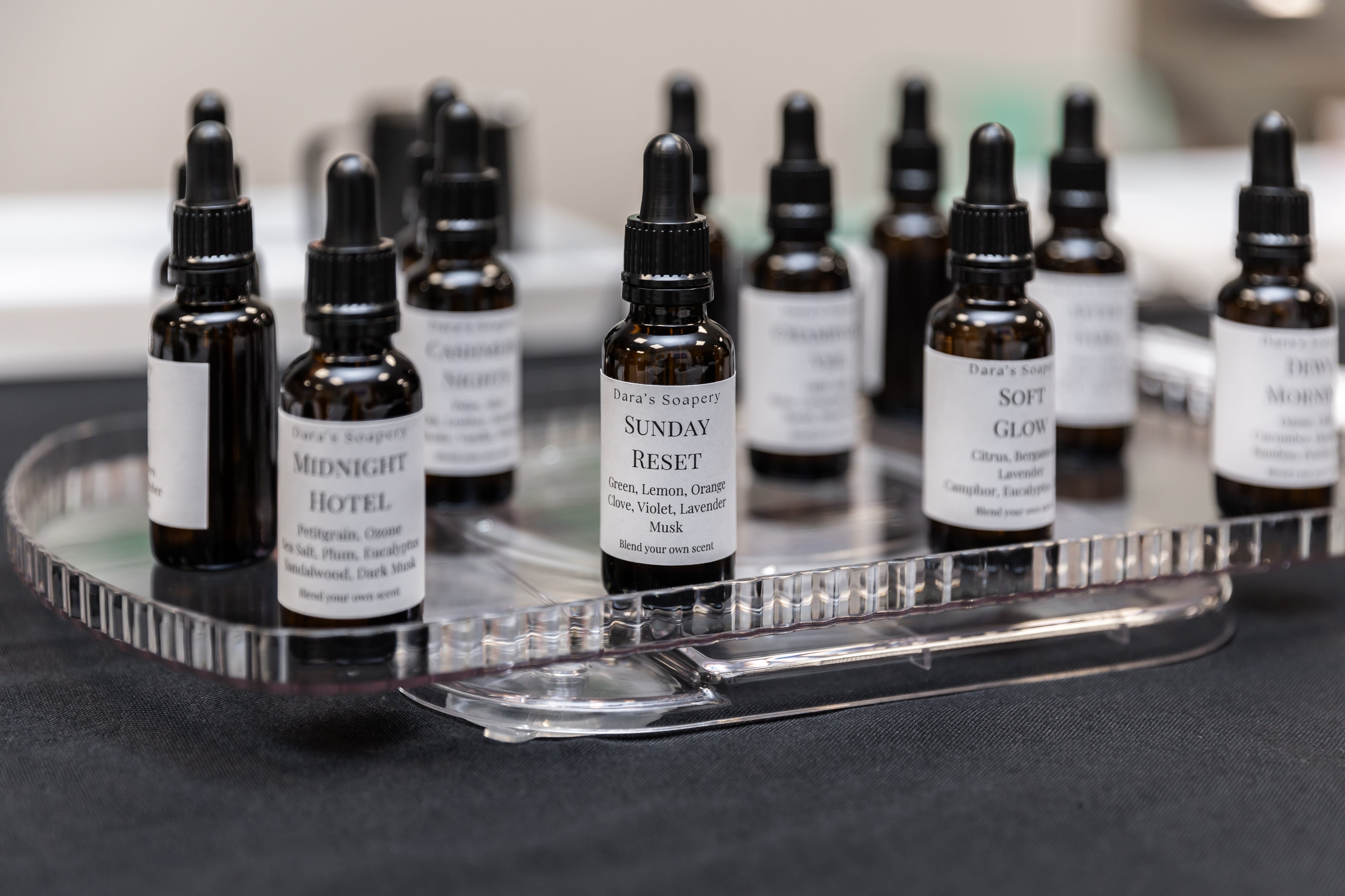 A group of small amber glass dropper bottles with white labels arranged on a clear acrylic tray. The labels show different product names including Midnight Hotel, Sunday Reset, Soft Glow, and others.