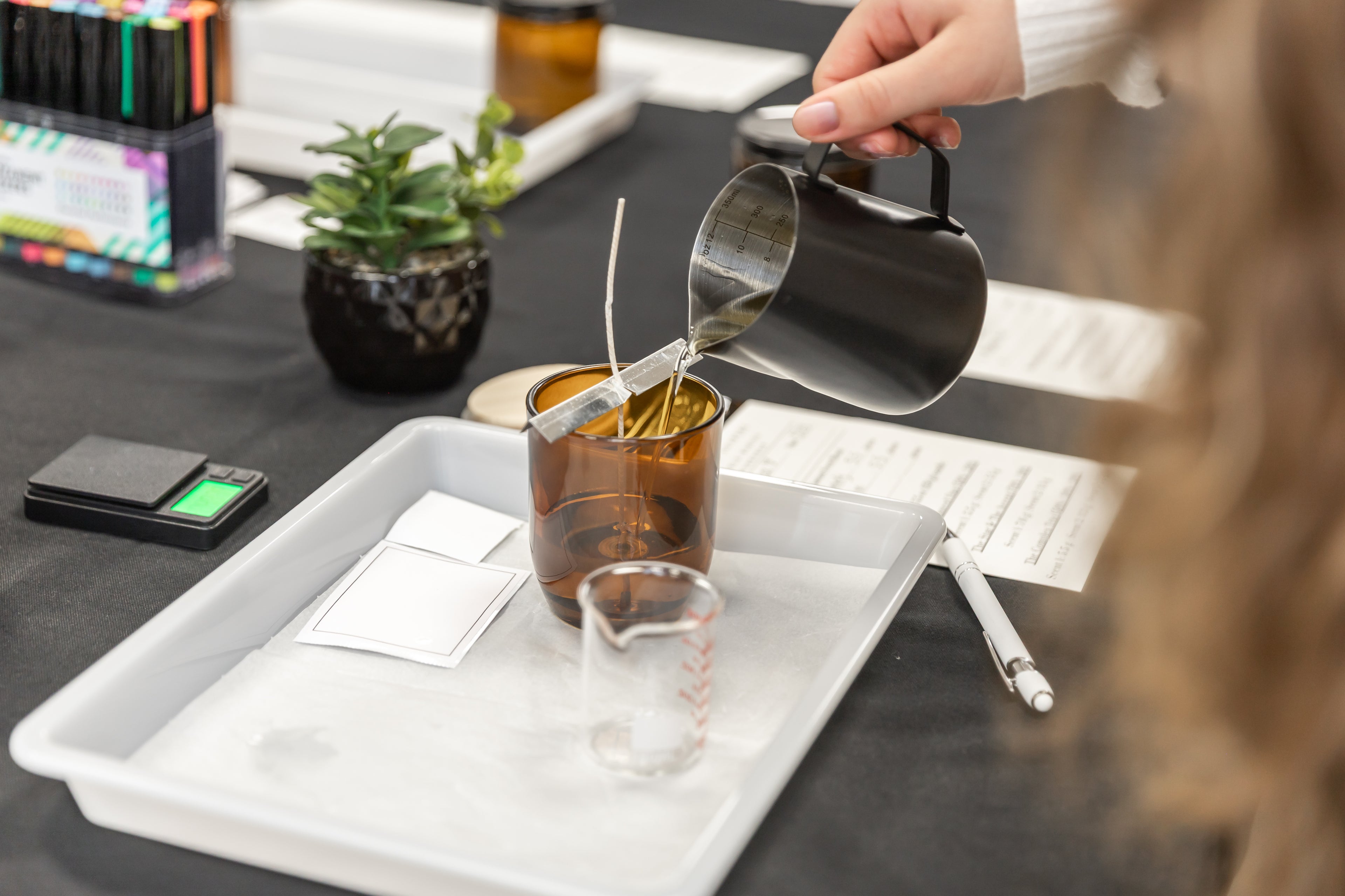 A person pouring hot wax from a metal pitcher into an amber glass container with wicks, set on a white tray at a craft or candle-making station.