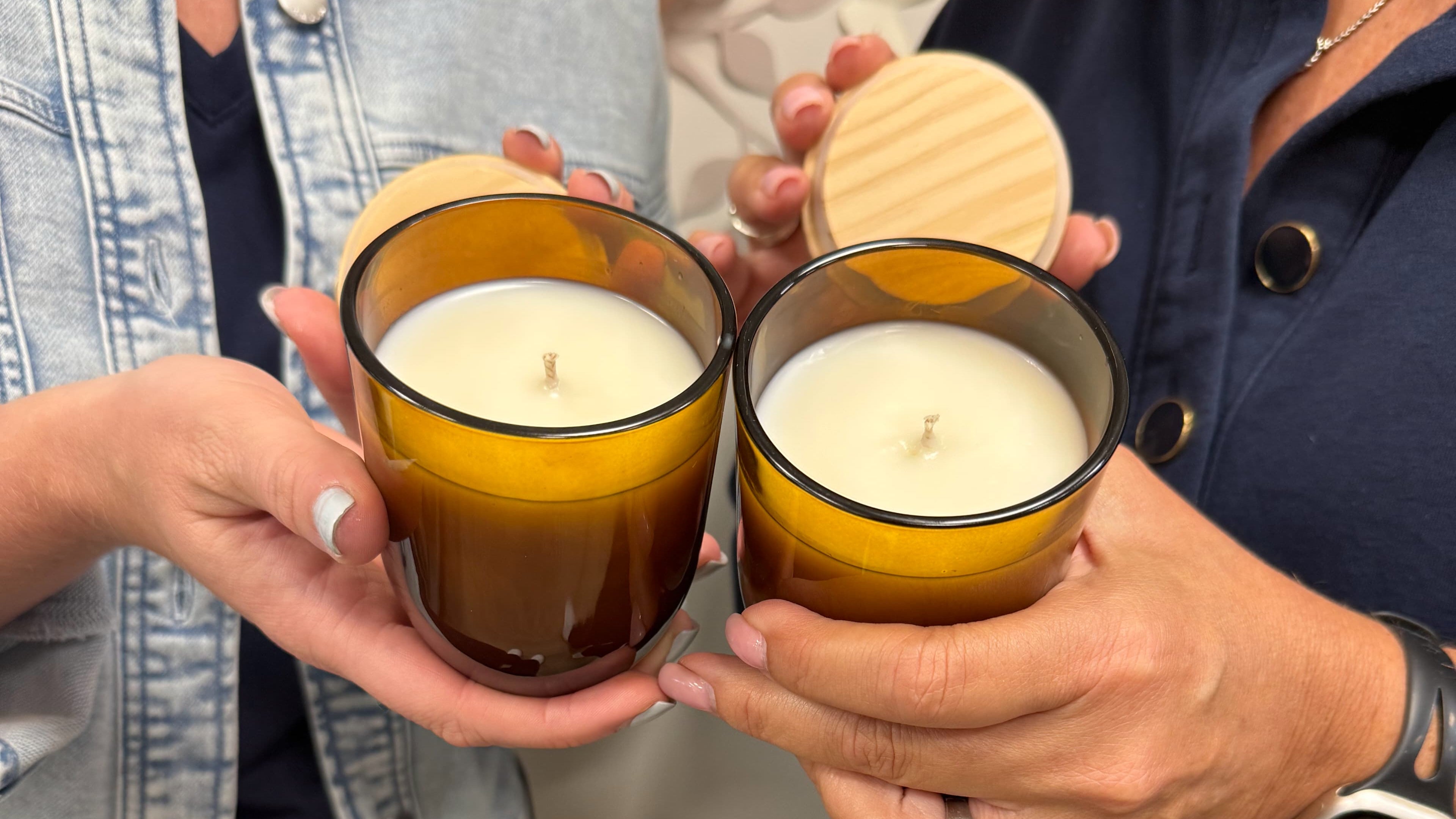 Two people holding amber glass jar candles with white wax and wooden lids in a cozy indoor setting.