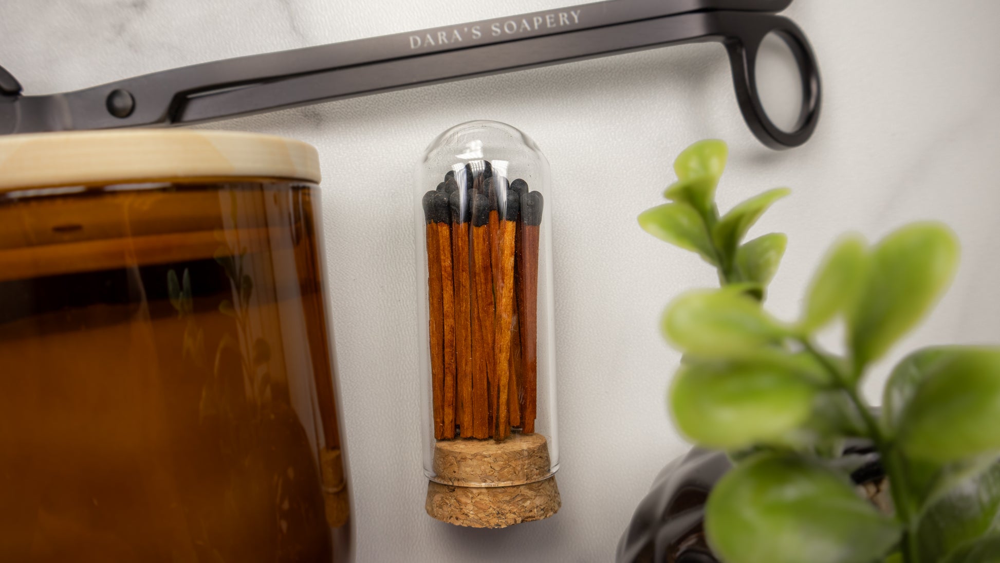 A small glass capsule vase filled with wooden matchsticks and black tips, mounted on a white wall. A large amber glass jar is partially visible on the left, and a green plant is blurred on the right. Included for candle club members at Dara's Soapery