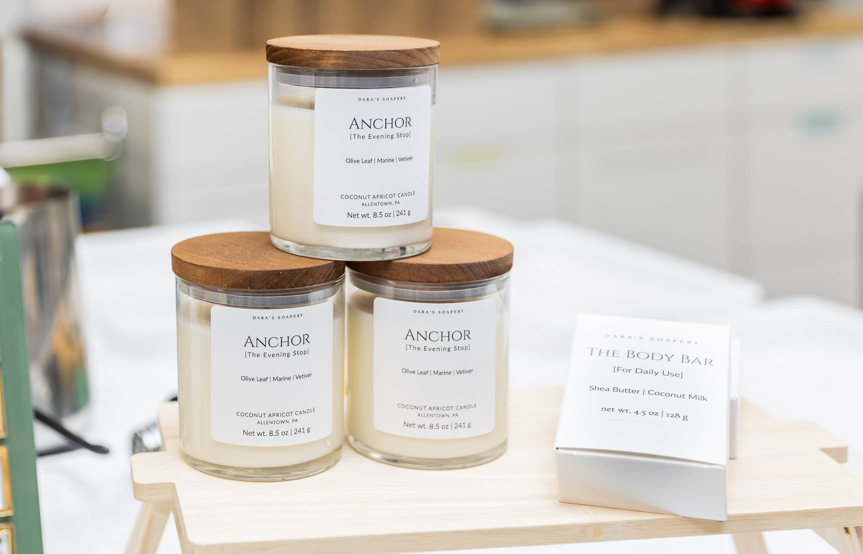 Three glass jar candles with wooden lids and white labels reading 'ANCHOR' are displayed on a small wooden stand in a bright indoor retail setting.