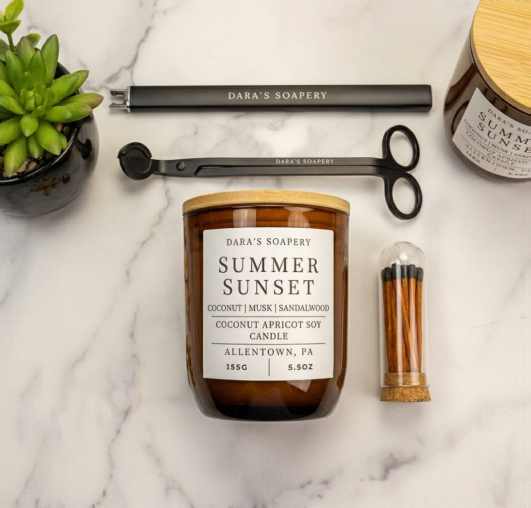 A styled flat-lay of a 'Summer Sunset' coconut apricot soy candle by Dara's Soapery, featuring an amber glass jar with a white label and a bamboo lid, surrounded by a black metal wick trimmer, a small succulent, and a match container on a white marble background.