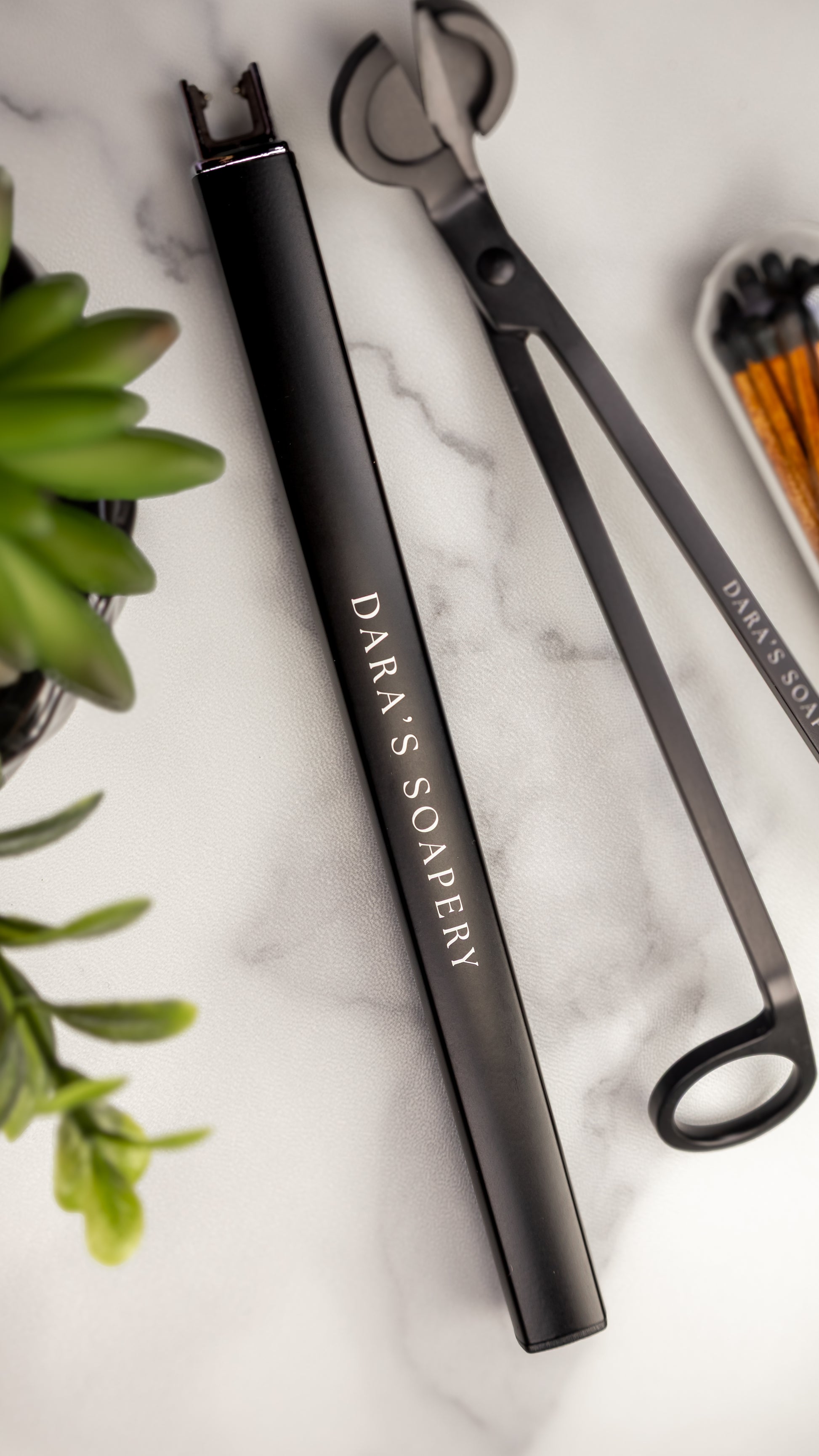 A black metal electric lighter with the brand name 'DARA'S SOAPERY' printed in white, arranged on a white marble surface next to a green plant. Included for members at Dara's Soapery. 
