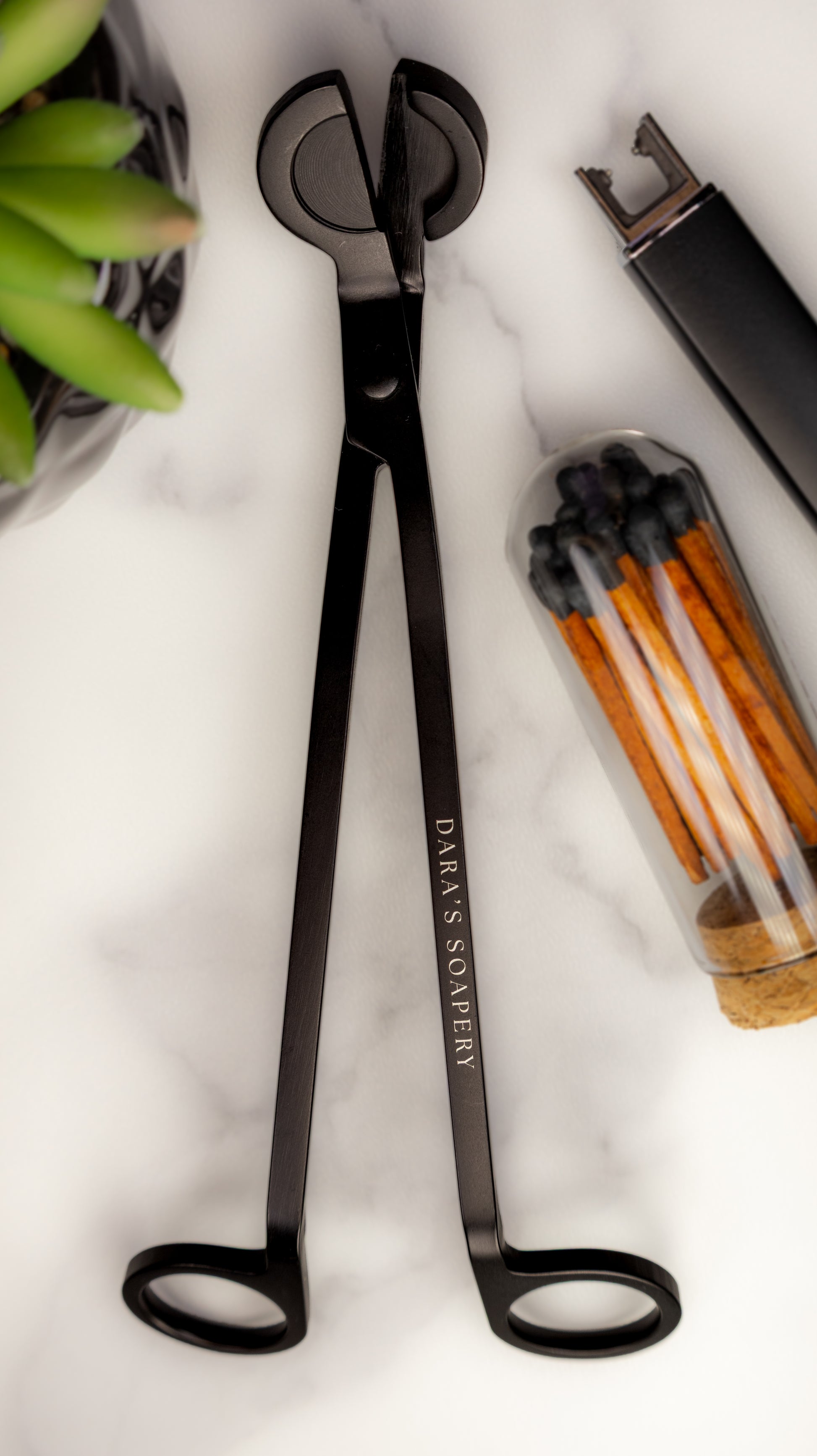 Black metal wick trimmer, shown on a white marble surface with decorative props including a plant, matches, and a lighter. Included for members at Dara's Soapery.
