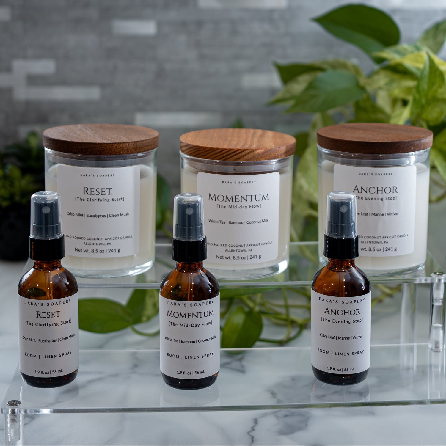 Set of candles and linen sprays including jars and bottles with wooden lids on a clear surface.