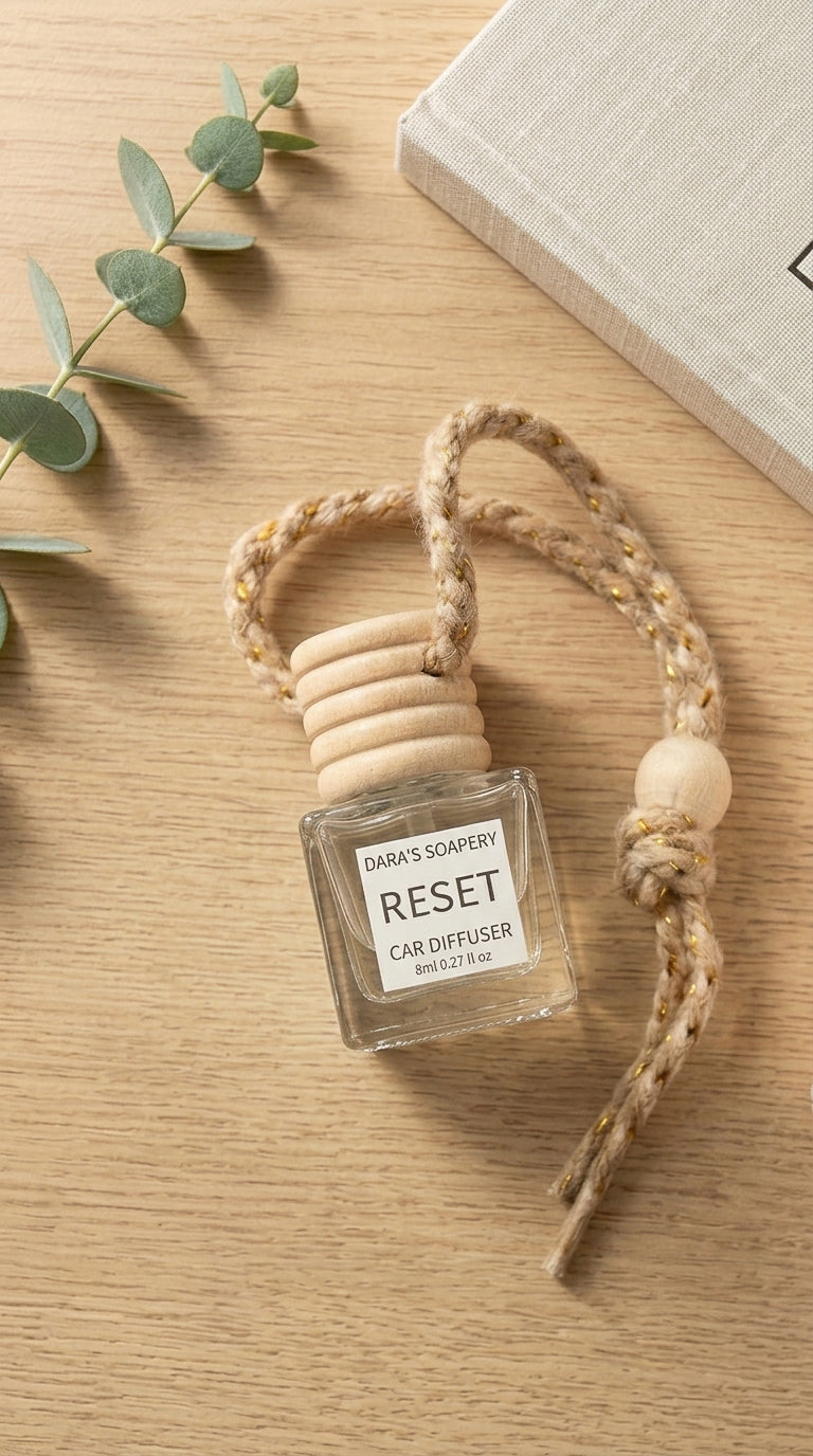 A clear glass car diffuser bottle with a beige rope loop and wooden cap, labeled 'DARAY'S SOAPERY RESET CAR DIFFUSER,' placed on a light wood surface with a book corner and eucalyptus leaves nearby.